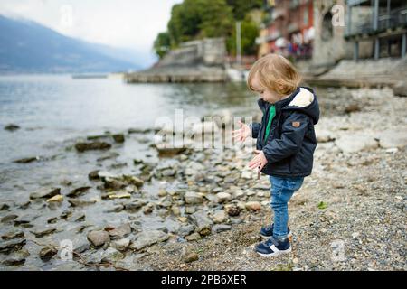 Süßer kleiner Junge, der im Hafen von Varenna, einer der malerischsten Städte am Ufer des Comer Sees, mit Kieselsteinen spielt. Varenna, Lombardei, Italien. Stockfoto