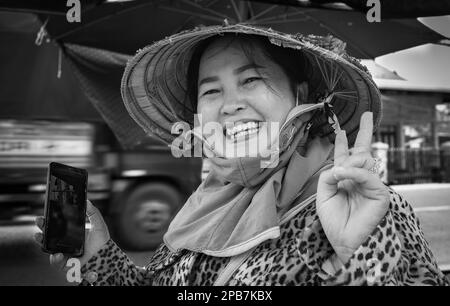 Eine vietnamesische Frau, die einen traditionellen konischen Hut trägt und ein Mobiltelefon in der Hand hält, lächelt und posiert für ein Foto in Tan Chau im Mekong-Delta in Vietnam Stockfoto