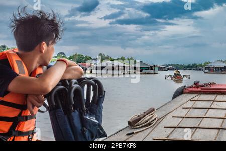 Ein junger Mann auf einem hölzernen Tender beobachtet ein traditionelles Fischerboot neben Fischfarmen auf dem Mekong bei Tan Chau in Vietnam. Stockfoto