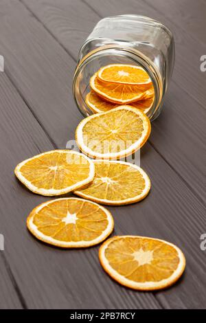 Trockene orangefarbene Scheiben werden aus einem Glasbehälter auf dem Tisch gestreut. Draufsicht. Dunkler Holzhintergrund. Stockfoto