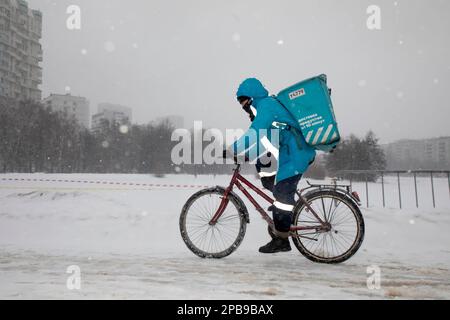 Moskau, Russland. 12. März 2023. Ein Kurier des Lieferservice Yandex Lavka fährt mit dem Fahrrad, um eine Bestellung an einen Kunden bei Schneefall in einem Wohngebiet in Moskau, Russland, zu liefern Stockfoto