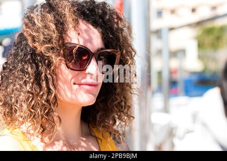 Nahaufnahme einer schönen jungen weißen Frau mit lockigem Haar und Sonnenbrille auf der Straße an einem sonnigen Tag. Stockfoto