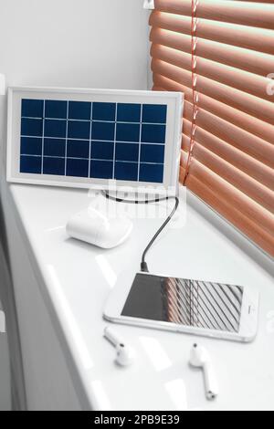 Tragbares Solarpaneel-Ladegerät, Mobiltelefon und Ohrhörer auf Fensterbank im Zimmer Stockfoto