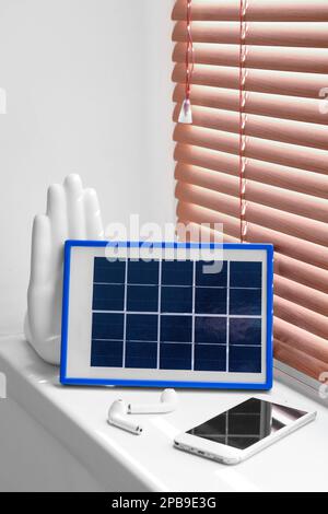 Tragbares Solarpaneel-Ladegerät, Mobiltelefon und Ohrhörer auf Fensterbank im Zimmer Stockfoto