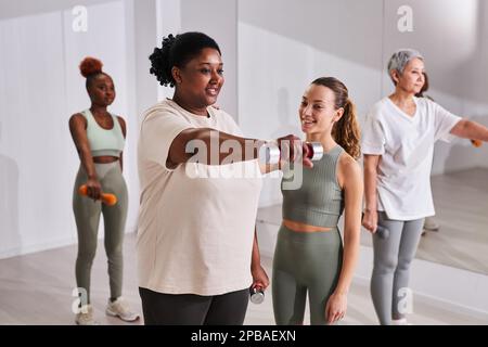 Eine afroamerikanische Übergewichtige trainiert mit einer Trainerin im Fitnessstudio, sie macht Bizeps-Übungen, indem sie Kurzhanteln hebt Stockfoto