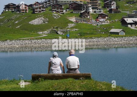 Ein älteres Ehepaar mit Blick nach hinten sitzt auf einer Bank am Melchsee in der Schweiz. Sie schauen auf das Wasser und auf die gegenüberliegende Seite des Sees zu Häusern. Stockfoto