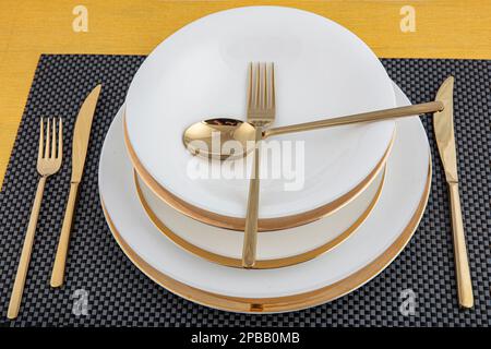 Teller und Utensilien, Teller, Schüssel und goldenes Besteck auf dem Esstisch, Seitenansicht. Moderne keramische Tischgeschirr, Besteck auf dem Esstisch. Stockfoto
