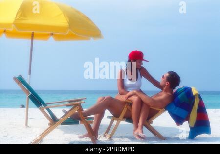 Ein jüngeres weißes Paar am Strand, wo eine Frau auf dem Schoß eines Mannes sitzt, während sie sich mit Liegestühlen und Sonnenschirmen im Sand am Meer unterhalten Stockfoto