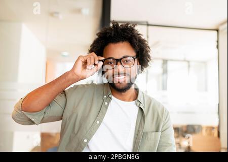 Attraktiver junger afroamerikanischer Geschäftsmann stilvolle Brille in Freizeitkleidung, die lächelt und in die Kamera schaut, ein Typ mit lockigem Afro-Haar, der in einem modernen Gemeinschaftsraum für Fotos posiert, passt die Brille an Stockfoto