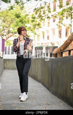 Porträt, hübsche junge asiatische Studentin in lässiger Kleidung und Gläsern, die mit ihrem Kaffee zum Mitnehmen durch das Campus-Gebäude spaziert Stockfoto