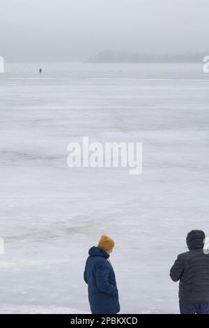 Ein paar Leute schauen vom Ufer auf einen Fischer auf dem dünnen Quelleis des Voronesch-Reservoirs. Jedes Jahr nehmen verzweifelte Liebhaber des Winterfischens das Eis des Voronesch-Reservoirs. Die Fischer werden nicht durch ein Auftauen oder sogar das Eintreffen des Frühlings aufgehalten. Die Aufregung treibt diese Menschen auf dünnem und manchmal verschwommenem Eis an, was zu Spannungen für den Dienst der Stadt führt. Rettungskräfte und Polizisten entfernen Fischer aus Eisschollen und fischen sie aus Gully, während medizinische Arbeiter um das Leben von Menschen kämpfen, die ertrinken oder kalt sind. Stockfoto