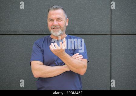 Selbstbewusster Mann mittleren Alters, der mit der Hand Gesten macht und die Kamera an einer grauen Wand lächelt Stockfoto