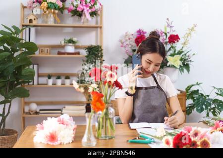 Floristen-Konzept, weibliche Floristin im Gespräch mit dem Kunden am Telefon und Notizen im Notebook im Geschäft. Stockfoto
