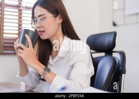 Geschäftskonzept, trinkt Geschäftsfrau Kaffee, bevor sie morgens im modernen Büro mit der Arbeit beginnt. Stockfoto