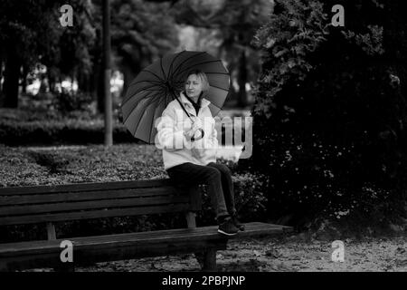 Eine Frau sitzt mit einem Regenschirm auf einer Parkbank. Schwarzweißfoto. Stockfoto