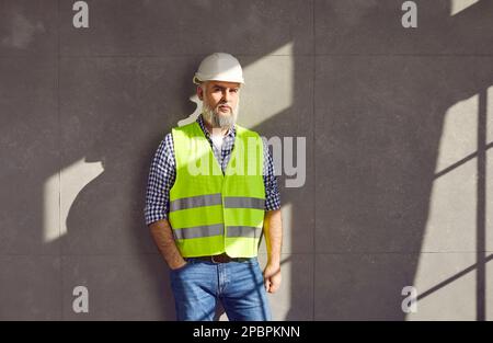 Reifer Baumeister oder Bauingenieur in Uniform und Schutzhelm, der an der grauen Hauswand steht Stockfoto