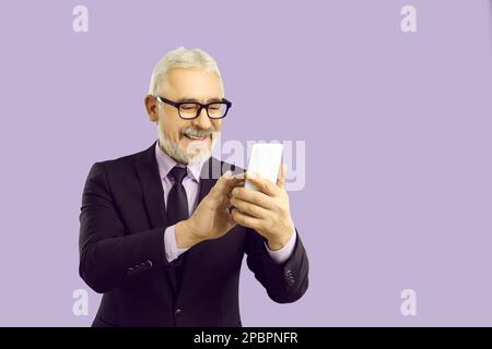Fröhlicher älterer Geschäftsmann im Anzug mit Smartphone, isoliert auf lila Hintergrund Stockfoto