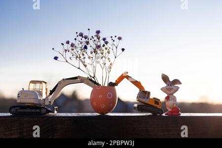 Zwei Spielzeugbagger-Modelle, eine Eierschale mit Blumen, ein Osterhasen im Hintergrund bei Sonnenuntergang. Osterferienkonzept für Bauleistungen Stockfoto