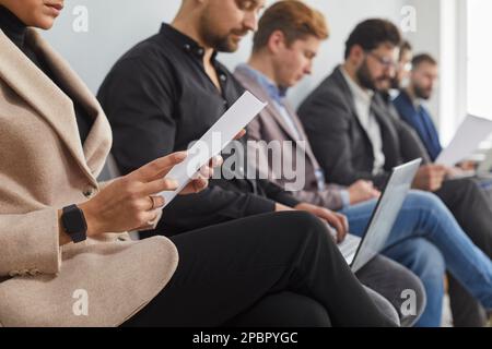 Die Gruppe der Kandidaten sitzt auf Stühlen in der Schlange und wartet auf ein Interview. Sie hält Fragebögen, Laptops und Mobiltelefone in der Hand. Stockfoto