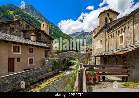 Der Alpenfluss fließt unter der mittelalterlichen Steinbrücke zwischen alten Häusern und der Pfarrkirche als Berge im Hintergrund im kleinen Dorf Chianale in Piemont, Stockfoto