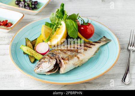 Gegrillte Meerbrasse mit Gemüse und Grün auf einem Holztisch Stockfoto