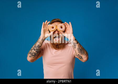 Aufgeregter, schreiender bärtiger Mann in Freizeitkleidung, der seine Augen mit Donuts bedeckt, isoliert auf blauem Hintergrund Stockfoto