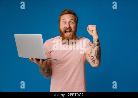 Ein bärtiger glücklicher Mann, der schreit und den Laptop hält, während er isoliert vor blauem Hintergrund steht Stockfoto
