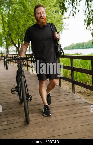 Ausgewachsener gutaussehender, tätowierter, bärtiger, ruhiger Mann in schwarzem Sportanzug, der zur Seite blickt, während er mit dem Fahrrad auf einer Holzbrücke im Park spaziert Stockfoto