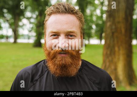 Porträt eines jungen, gutaussehenden, lächelnden Rothaarigen, der in die Kamera schaut, während er im grünen Park steht Stockfoto
