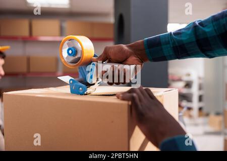 afroamerikanische Männer halten Packbandpistole und versiegelten Pappkarton. Lagerarbeiter schließt das Paket mit Klebstoff und bereitet den Kundenauftrag für den Versand aus nächster Nähe vor Stockfoto
