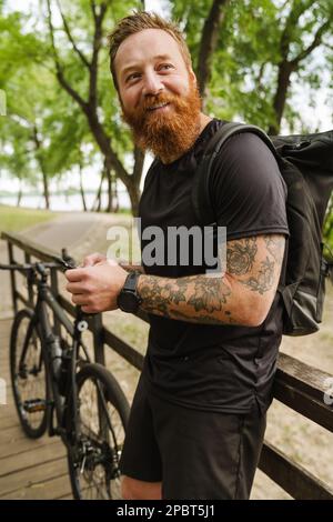 Porträt eines jungen, gutaussehenden, tätowierten, bärtigen Mannes, der das Telefon hält und zur Seite schaut, während er sich in der Sonne neben dem Fahrrad auf einem Holzgeländer lehnt Stockfoto