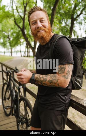 Porträt eines jungen, gutaussehenden, tätowierten bärtigen Mannes, der sein Handy in der Hand hält und in die Kamera schaut, während er sich auf ein Holzgeländer lehnt und neben dem Fahrrad steht Stockfoto