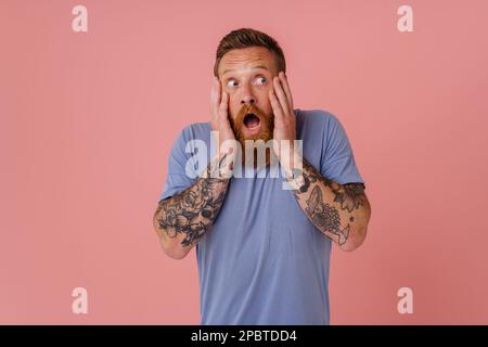 Erwachsener, mit einem rothaarigen Bart tätowierter, schockierter Mann im T-Shirt, der seine Wangen hält und zur Seite schaut, während er über einem isolierten pinken Hintergrund steht Stockfoto