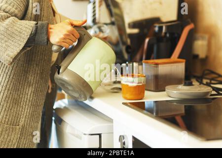 Eine Frau, die Wasser aus dem Wasserkocher in den Becher am Tisch in der Küche gießt. Stockfoto