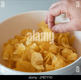 Die Hand eines Mannes greift nach Kartoffelchips in einer weißen Keramikschüssel Stockfoto