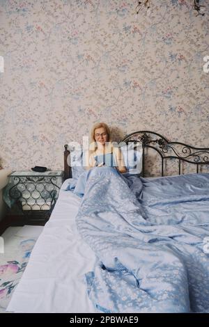 Frau mittleren Alters, die sich im Bett ausstreckt, alleine aufwacht und eine Tablette in der Hand hält Stockfoto