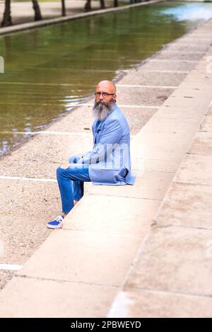 Ein Mann mittleren Alters, kahl, mit Bart, der in einem Park sitzt und sich entspannt Stockfoto