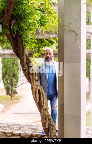 Lateinamerikanischer, kahler, bärtiger Mann, der hinter einem Baum in einem Park steht. Stockfoto