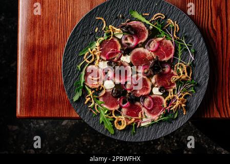Marmorfleisch-Carpaccio mit Soße und Kräutern auf einem schwarzen Teller Stockfoto