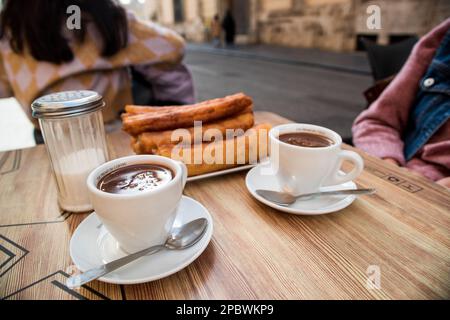 Churros und zwei Tassen Schokolade auf dem Terrassentisch, fertig zum Essen. Stockfoto