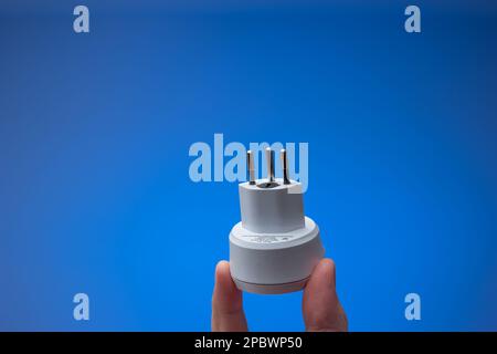 Steckdosenadapter in Schweizer Ausführung, der mit dem Stecker in der Hand gehalten wird. Nahaufnahme im Studiostil, isoliert auf blauem Hintergrund. Stockfoto