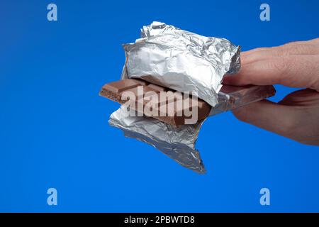 Generischer Schokoriegel in Aluminiumfolie, der von männlichen Händen gehalten wird. Nahaufnahme des Studios, isoliert auf blauem Hintergrund. Stockfoto