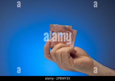 Papierbanknote mit einem kanadischen 50-Dollar-Schein, zwischen den Fingern gehalten von kaukasischer männlicher Hand. Nahaufnahme des Studios, isoliert auf blauem Hintergrund. Stockfoto