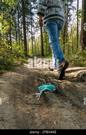 Medizinische Gesichtsmaske verstreut in der Natur.nicht wiedererkennbarer Mensch geht weg, nachdem er eine Maske auf den Boden auf einem Fußweg im Wald fallen ließ. Niedriger Weitwinkelstreuer Stockfoto