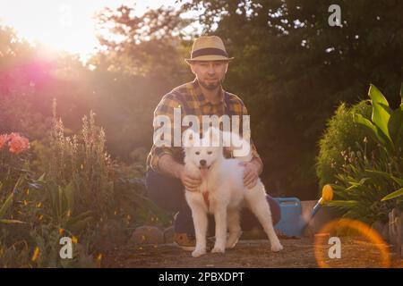 Ein hübscher Weißer in legerer Kleidung, der mit einem weißen Samoyerten Hündchen posiert. Sonniger Garten im Hintergrund. Das Konzept des Trainings von Haustier und Happy Do Stockfoto