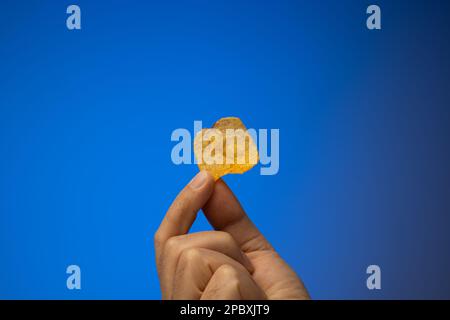 Ein Kartoffelchip, der von einem weißen Mann in der Hand gehalten wird. Nahaufnahme des Studios, isoliert auf blauem Hintergrund. Stockfoto