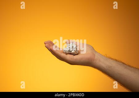 Die Zinnfolie in der Küche wurde zu einem Ball zerbrochen, der von einem weißen Mann in der Hand gehalten wurde. Nahaufnahme des Studios, isoliert auf orangefarbenem Hintergrund. Stockfoto