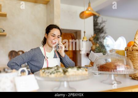 Glückliche junge Frau in der Schürze, Start-up wird wahr. Konzept für kleine Unternehmen. Familiengeschäft, Café-Laden. Telefonieren, Befehle entgegennehmen. Stockfoto