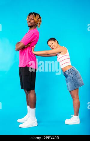 Seitenansicht einer positiven jungen ethnischen Frau mit kurzen dunklen Haaren in legerer Kleidung, die lächelt und einen großen afroamerikanischen Freund gegen Blue BA wegschiebt Stockfoto