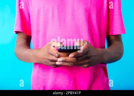 Ein unerkannter afroamerikanischer Typ in einem rosa T-Shirt, der vor blauem Hintergrund auf seinem Handy surft Stockfoto